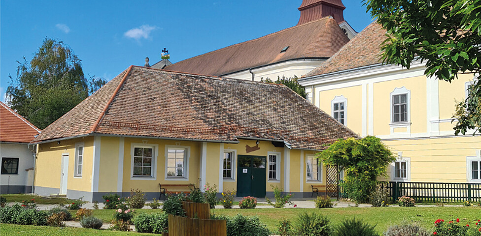 Innenhof Bauernhof Unternalb. Freistehendes kleines Haus mit Spitzdach und braunen Ziegeln gedeckt. Fassadenfarbe in gelb. Im Vordergrund sieht man Grasfläche mit einer Kräuterschnecke aus Holz. Im Hintergrund sieht man das Kirchturmdach der Kirche.