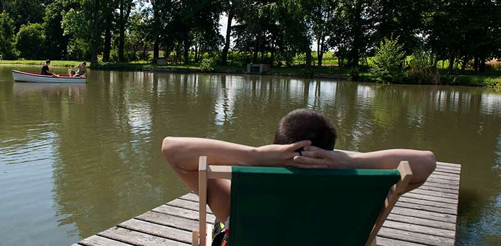 Person sitzt in Liegestuhl aus Holz. Ansicht von hinten. Der Liegestuhl steht am Steg des Teiches. Links hinten im Wasser Ruderboot mit 2 Personen.Im Hintergrund sieht man das Wasser des Teiches und hohe dunkelgrüne Bäume, die das Ufer säumen.